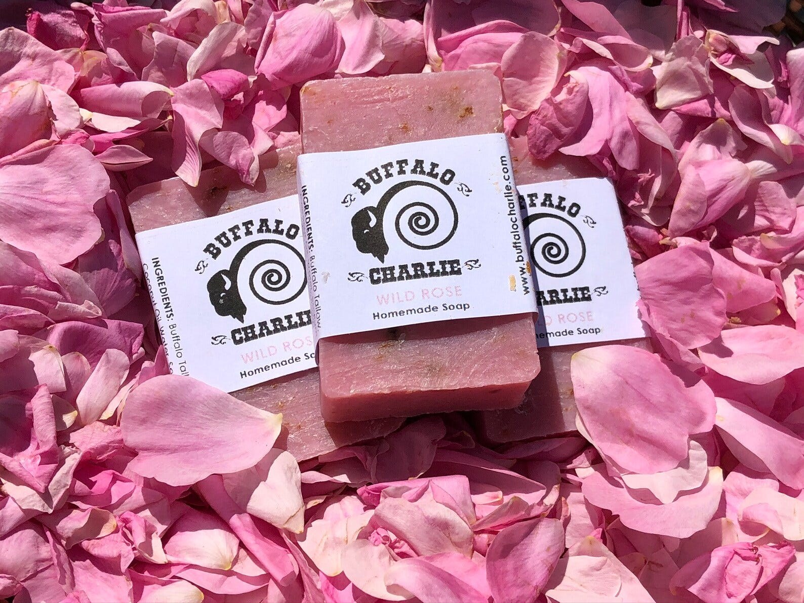 Three bars of pale pink Wild Rose soap in Buffalo Charlie wrappers, resting on a bed of wild rose petals